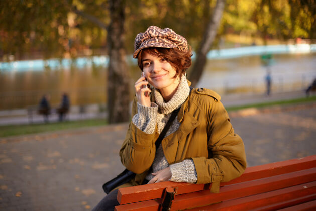 休闲可爱的年轻积极的棕色头发女性 随意的发型 坐在城市公园的木凳上愉快地微笑着 愉快地打电话衣服针织公园