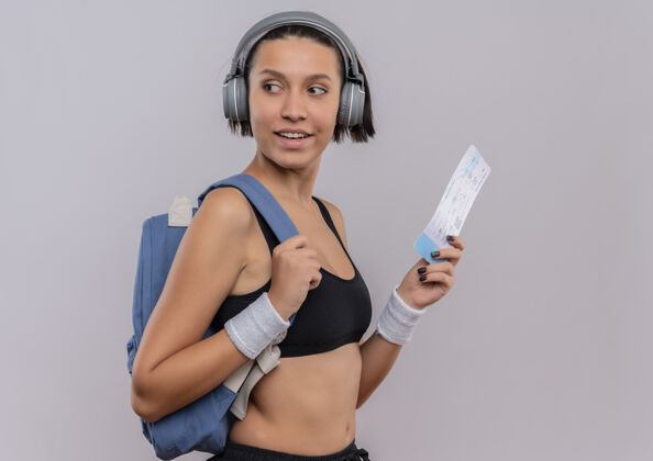 运动员身穿运动服 头戴耳机 手持机票 面带微笑地站在白墙上的年轻健身女士头看健身