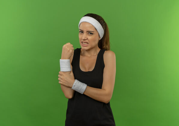 运动员穿着运动服的年轻健身女 头箍碰到手腕 站在绿墙上疼痛难忍疼痛运动站立
