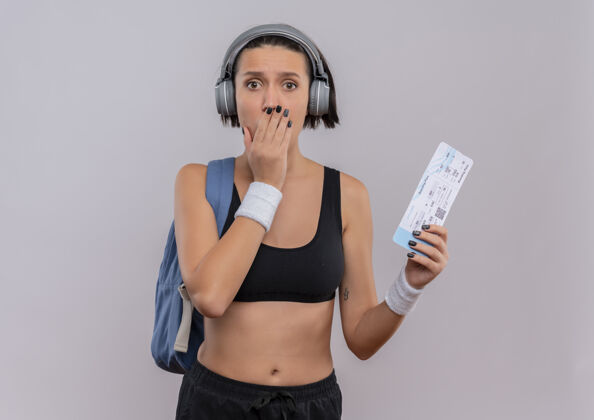 封面穿着运动服的年轻健身女士头戴耳机背着背包拿着机票手捂着嘴站在白墙上公民震惊健身