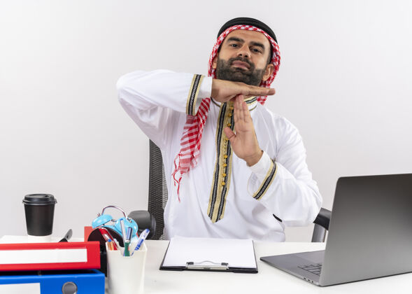 工作穿着传统服装的阿拉伯商人坐在桌旁 拿着笔记本电脑 在办公室里用手做着暂停的手势制造商务人士坐着