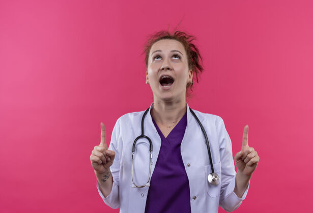 退出年轻的女医生穿着白大褂 带着听诊器 用食指站在粉红色的墙上 兴奋而惊讶地指着我惊喜目录外套