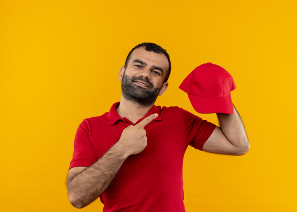 胡子一个留着胡须 身穿红色制服 手拿帽子 手指指着帽子的送货员站在橙色的墙上 自信地微笑着男人指着手指