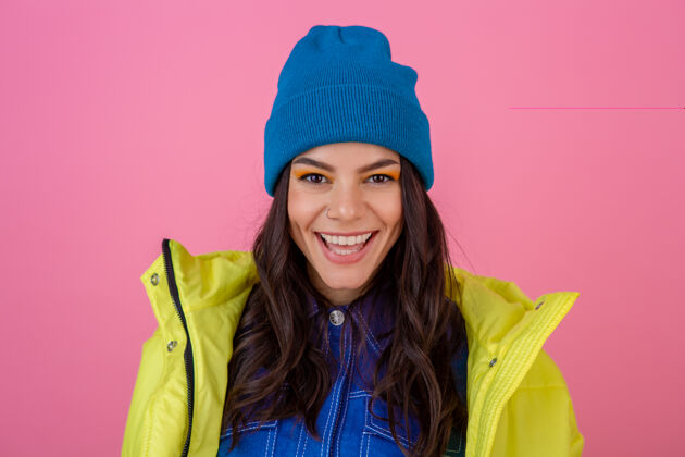 快乐迷人微笑的时尚女性的肖像在冬天的时尚造型在粉红色的墙上穿着鲜艳的霓虹黄夹克 戴着蓝色的针织帽子 穿着暖和的衣服温暖微笑针织