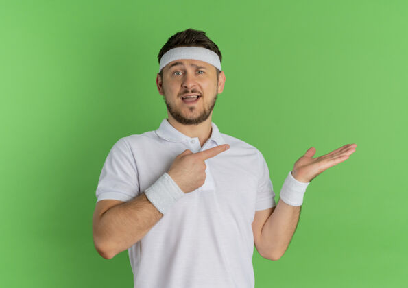 侧线一位身穿白衬衫的年轻健身先生 头上戴着一条指环 手指指向一边 他的手臂站在绿色的墙上人运动手臂