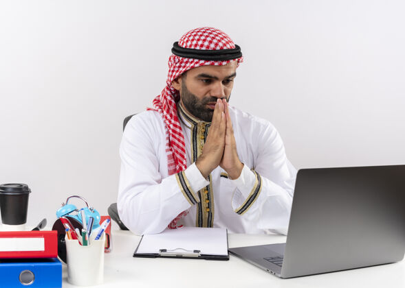 手身着传统服装的阿拉伯商人手牵手坐在办公桌旁用笔记本电脑办公希望商务人士坐