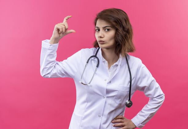 支架身穿白大褂的年轻女医生带着听诊器 用手指显示小尺寸标志 测量标志站在粉红色的墙上符号显示外套