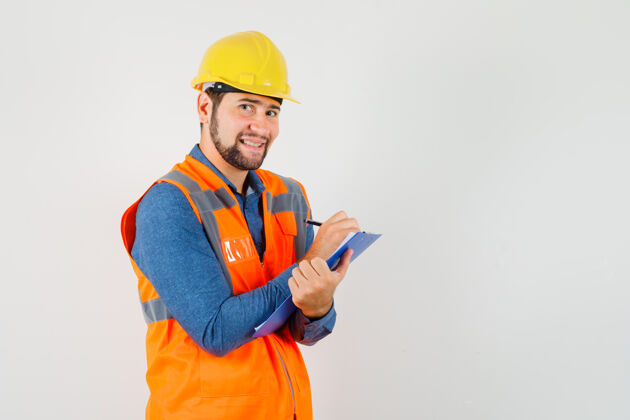 工匠年轻的建筑工人穿着衬衫 背心 头盔在剪贴板上做笔记 看上去很高兴正视图制服注意项目