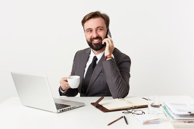 衬衫摄影棚拍摄的是一位留着胡须 留着短发的男性 他正坐在工作桌前 端着一杯咖啡 用智能手机打电话 脸上露出灿烂的笑容爱未剃须理发