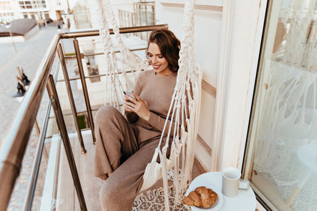卷发在阳台吃午饭时 可爱的女孩在发短信优雅的年轻女子坐在阳台上 手里拿着牛角面包和茶手机思想户外