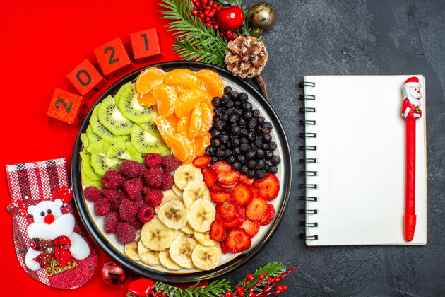 风景晚餐盘上收集新鲜水果的俯视图装饰配件杉木树枝和数字圣诞袜放在红色餐巾上下一个笔记本用黑色背景上的钢笔笔记本树枝蔬菜