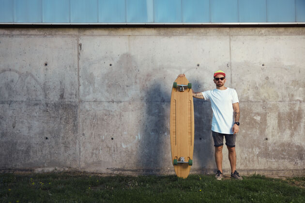 享受严肃的冲浪者 胡子 纹身和太阳镜站在他的长板旁边灰色滑板长板