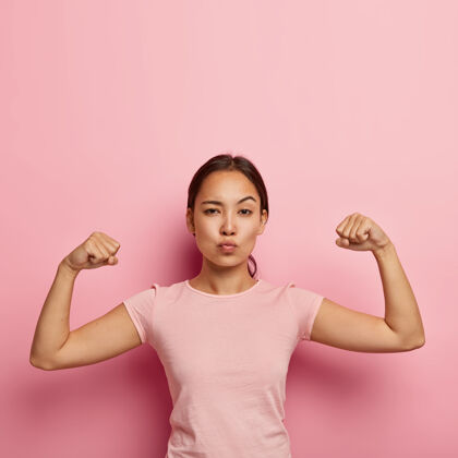 手势自信严肃的亚洲女孩的照片保持嘴唇合拢 显示她的肌肉和力量 不化妆 穿着休闲t恤 隔离在粉红色墙上 上面有复制空间供参考模特肌肉休闲