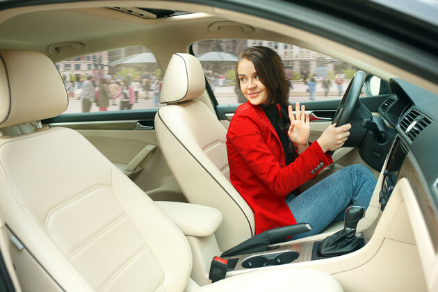 汽车在城市里开车年轻漂亮的女人开着车年轻漂亮的白人模特穿着优雅时尚的红色夹克坐在现代汽车内饰上旅行乘客司机