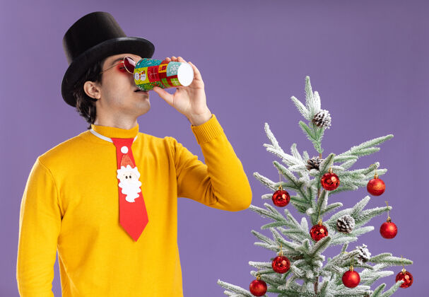 穿着身穿黄色高领毛衣 戴着黑色帽子和滑稽领带的年轻人站在紫色墙上的圣诞树旁 从五颜六色的纸杯里喝水杯子男人领带