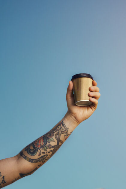 人纹身的白人高举一个米色一次性咖啡杯 映衬着湛蓝的天空饮料棕色早晨