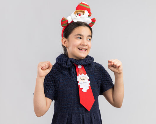 有趣小女孩穿着针织连衣裙 头上系着一条红色领带 头上戴着有趣的圣诞戒指 握紧拳头 快乐而兴奋快乐快乐圣诞快乐