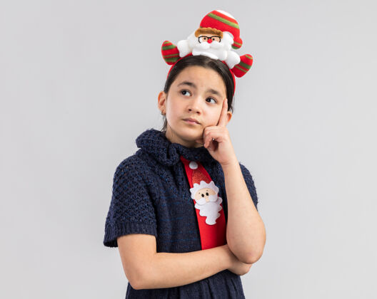 思想小女孩穿着针织连衣裙 头上系着一条红色领带 头上戴着一个滑稽的圣诞边 神情沉思地抬头思考年圣诞快乐穿着