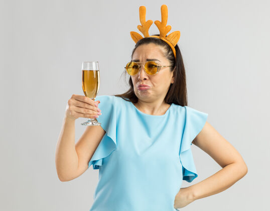 香槟身穿蓝色上衣的年轻女子戴着滑稽的鹿角边 戴着黄色的眼镜 手里拿着一杯香槟 困惑地 不高兴地看着它圣诞快乐黄色顶