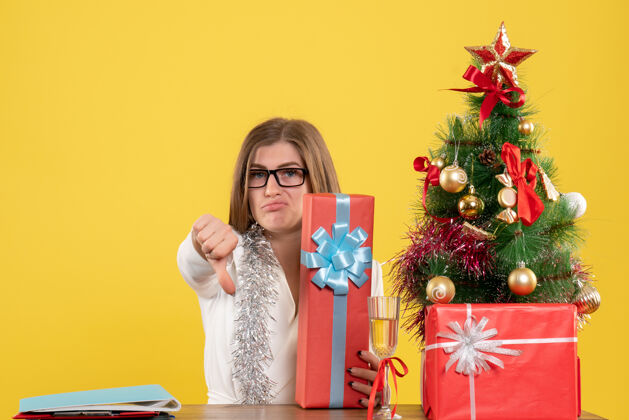 包正面图：女医生坐在桌子前 拿着礼物 黄色背景上的圣诞树和礼盒背景坐快乐