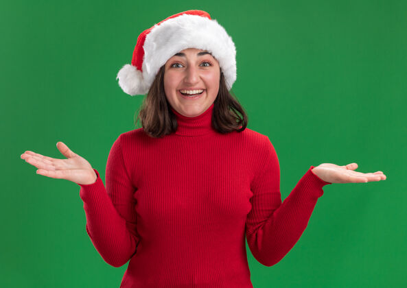 年轻人穿着红色毛衣 戴着圣诞帽的年轻女孩看着镜头 脸上洋溢着幸福的表情 双臂伸向两侧 站在绿色的背景上圣诞老人站帽子