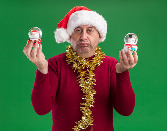 穿着中年男子戴着圣诞老人帽 脖子上戴着金属丝 手里拿着圣诞雪球 表情怀疑地球仪举行怀疑