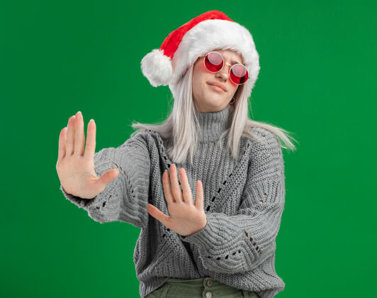 毛衣年轻的金发女郎穿着冬季毛衣 戴着圣诞老人帽 戴着红色眼镜 看起来很不高兴 双手站在绿色背景上做着防御手势年轻站着眼镜