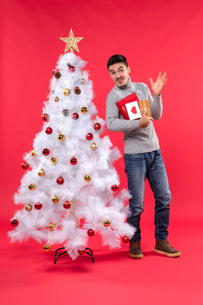 年轻人英俊有趣的年轻人站在装饰过的白色新年树旁 手里拿着他的礼物圣诞树男人年轻人