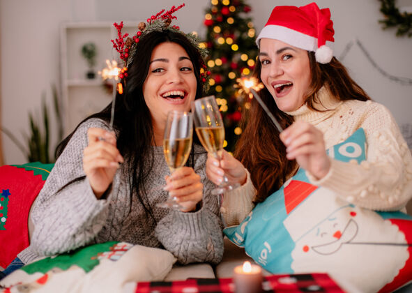 快乐快乐的年轻女孩戴着圣诞帽 碰杯香槟 拿着火花灯坐在扶手椅上 在家里享受圣诞时光圣诞快乐坐着漂亮