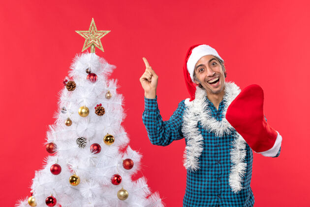 圣诞树一个微笑的年轻人戴着圣诞老人的帽子 穿着一件蓝色的衬衫 穿着他的圣诞袜子礼物庆祝衬衫