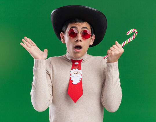 帽子一个戴着帽子 戴着圣诞领带 戴着眼镜 手里拿着圣诞糖果 手被隔离在绿墙上的年轻人吓坏了穿着害怕圣诞节