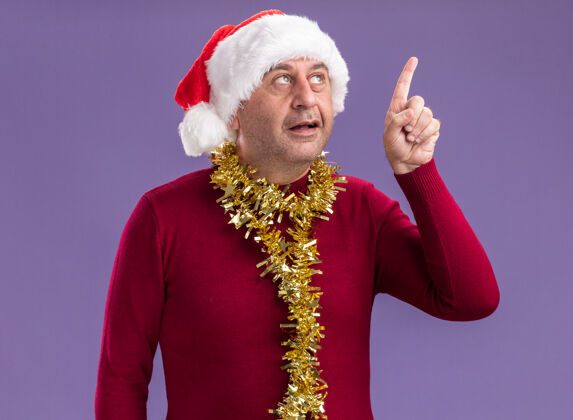 微笑中年男子戴着圣诞圣诞老人帽子 脖子上戴着金属丝 脸上露出微笑 食指站在紫色的背景上有了新的想法脸指数聪明