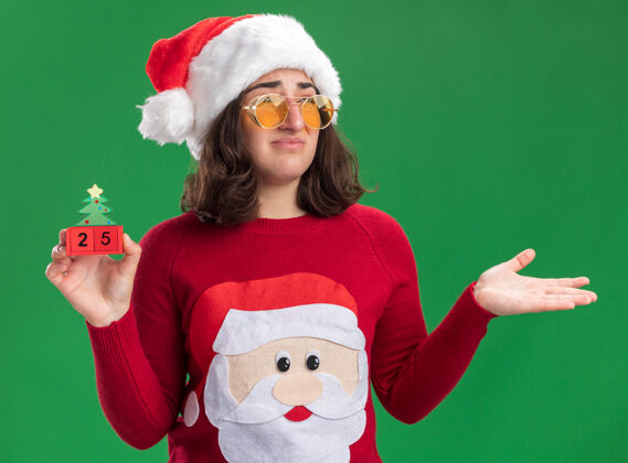 二十穿着圣诞毛衣的小女孩戴着圣诞帽 戴着眼镜 手里拿着25号的玩具立方体 困惑地站在绿色的墙上 抬起胳膊戴着毛衣数字