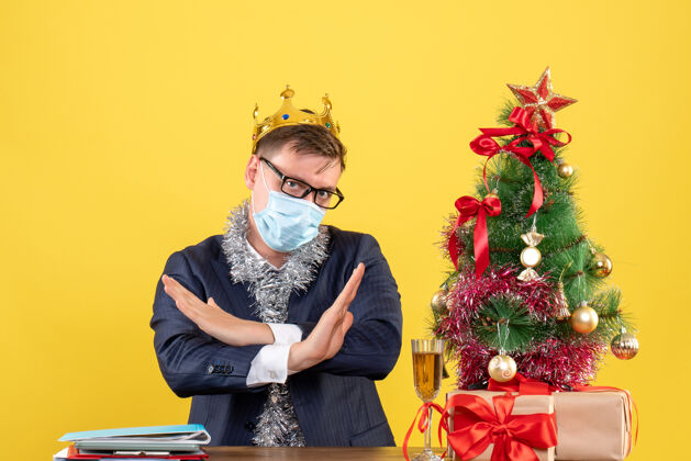 颈部支架商务人士交叉双手坐在圣诞树旁的桌子前 黄色墙壁上的礼物支持支架面具
