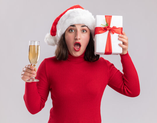 站着穿着红色毛衣 戴着圣诞帽的年轻女孩站在白色的墙上 手里拿着一杯香槟和一份礼物玻璃杯圣诞节毛衣