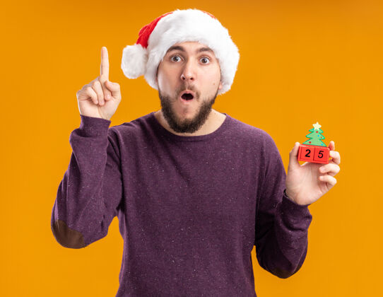 索引穿着紫色毛衣 戴着圣诞帽的年轻人展示着25号玩具立方体 惊讶地展示着食指站在橙色的墙上圣诞节毛衣二十