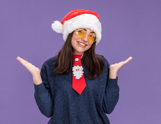 领带微笑的年轻白人女孩戴着太阳眼镜 戴着圣诞帽 打着圣诞领带 双手张开 在紫色的背景下 有着独立的复制空间快乐背景圣诞老人
