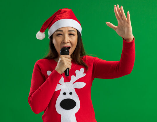 年轻人戴着圣诞老人帽 穿着红色毛衣的年轻女子手持麦克风快乐地唱着歌 站在绿色的背景下离开了圣诞老人圣诞节退出