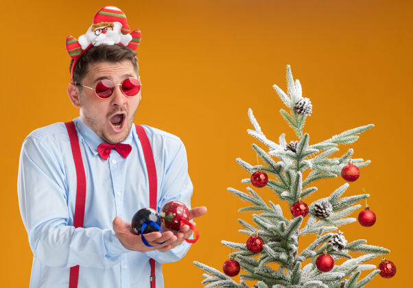 男人戴着吊带领结的年轻人站在圣诞树旁 戴着圣诞老人和红眼镜 神情迷茫和惊讶 手里拿着橘色墙上的圣诞树玩具吊带年轻困惑