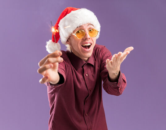 圣诞老人令人印象深刻的年轻金发男子戴圣诞帽和眼镜伸出节日火花显示空手孤立在紫色墙上显示穿着走向