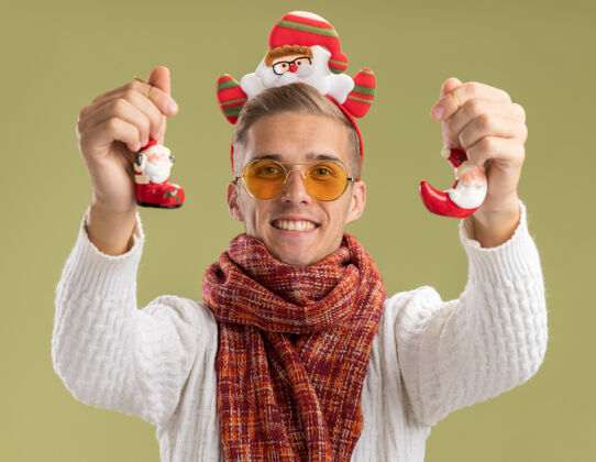 帅气快乐的年轻帅哥戴着圣诞老人的头带和围巾 向孤零零的橄榄绿墙上伸出圣诞老人的圣诞饰品戴着年轻圣诞