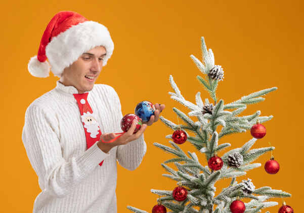 圣诞老人快乐的年轻帅哥戴着圣诞帽 打着圣诞老人的领带 站在装饰好的圣诞树旁 拿着圣诞球 看着橙色墙上孤立的装饰品圣诞节近球