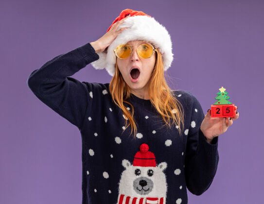 手害怕的年轻漂亮女孩穿着圣诞毛衣和帽子戴着眼镜拿着圣诞玩具把手放在头上孤立在紫色背景上害怕女孩帽子