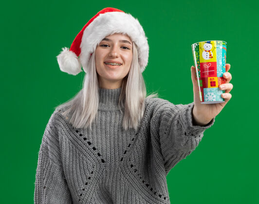圣诞老人年轻的金发女郎穿着冬衣 戴着圣诞帽 戴着一个漂亮的纸杯 站在绿色的背景下 面带微笑地看着摄像机金发杯子脸