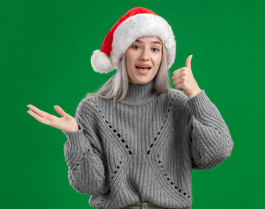 站着年轻的金发女郎穿着冬季毛衣 戴着圣诞帽 看着相机 快乐而愉快地竖起大拇指 在绿色的背景上 伸出手臂展示复制空间帽子年轻毛衣