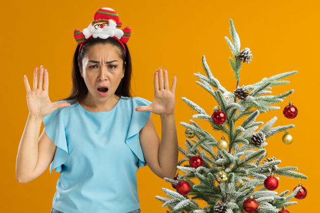 举起身穿蓝色上衣的年轻女子头戴滑稽的圣诞环 看着摄像机 愤怒的脸上举着双臂 站在橙色背景下的圣诞树旁大喊大叫脸年轻喊叫