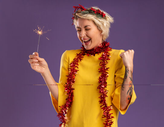 手势快乐的年轻金发女人戴着圣诞花环 脖子上戴着金箔花环 手里拿着节日火花灯 做着“是”的手势 闭着眼睛孤立地站在紫色的墙上头脖子年轻