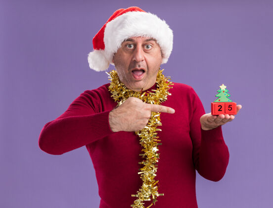 圣诞节中年男子戴着圣诞老人的帽子 脖子上戴着金属丝 手里拿着玩具方块 上面写着25岁的日期 用食指指着他们 站在紫色的背景下惊讶地看着他们帽子指数脖子