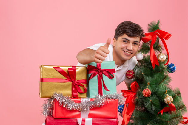 快乐圣诞礼物和粉红色墙上的圣诞树周围的年轻人的正面视图新郎丝带节日
