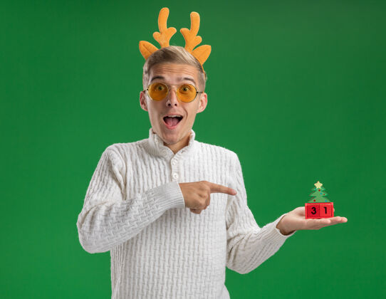 英俊给人印象深刻的年轻帅哥戴着驯鹿鹿角头带戴着眼镜拿着并指着圣诞树玩具和日期看着隔离在绿色墙上的一面印象深刻日期头带